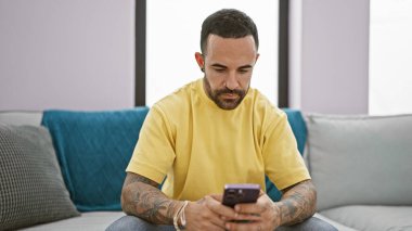 Evde akıllı telefon kullanan sakallı yakışıklı bir İspanyol adam. Sıradan giyinmiş ve dövmeli..