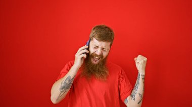 Aşırı neşeli genç kızıl saçlı adam, izole edilmiş bir kırmızı arka planda telefonla konuşurken kendinden geçmiş bir şekilde kutlama yapıyor.