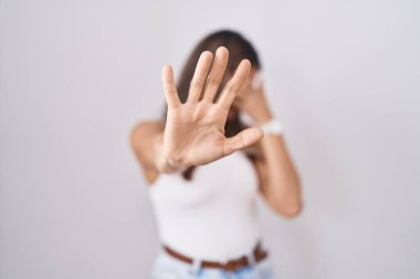 Latin kökenli genç bir kadın beyaz arka planda duruyor. Gözleri ellerle kaplıyor ve üzgün ve korku dolu bir ifadeyle dur hareketi yapıyor. utangaç ve olumsuz kavram. 