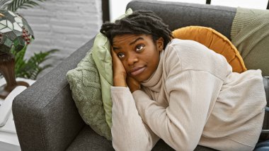 Düşünceli, genç, siyah bir kadın. Rastalı koltuklarda uzanmış, rahat yastıklarla çevrili..