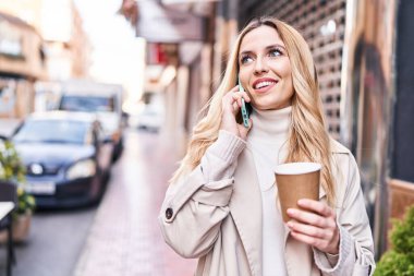 Young blonde woman talking on smartphone drinking coffee at street