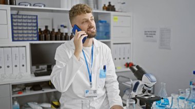 Sakallı, mavi gözlü, laboratuvar önlüğü giyen ve telefonla konuşan genç, beyaz bir adam bilimsel ekipmanlarla dolu bir laboratuarda duruyor..