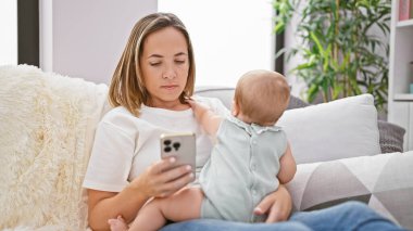 Rahat anne, kanepede oturuyor, akıllı telefonuyla mesajlaşırken ciddi bir ifade takınıyor, küçük kızıyla evde güzel bir anı paylaşıyor.