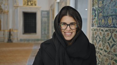 İstanbul 'un tarihi sarayında gözlük ve siyah başörtüsü takan gülümseyen bir kadın süslü fayansların önünde duruyor..