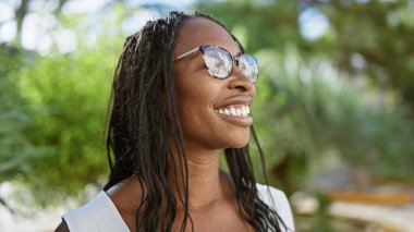 Kıvırcık saçlı, güneşli bir parkta gözlük takan mutlu Afrikalı Amerikalı kadın.