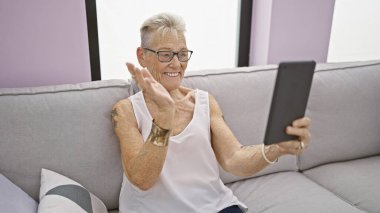 Neşeli, gri saçlı, son sınıf öğrencisi bir kadın evdeki kanepede oturup rahatlarken eğlenceli bir video görüşmesi yapıyor. Sıcak bir gülümsemeyle mutluluğu ifade ediyor.