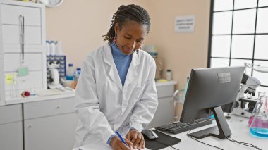 Kapalı bir laboratuvarda çalışan Afrikalı bir Amerikalı kadın bilim adamı bilgisayardaki verileri analiz ediyor..