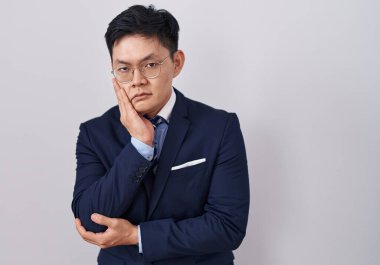 Genç Asyalı takım elbiseli ve kravatlı bir adam kollarını kavuşturarak depresyondan sıkılmış ve yorgun göründüğünü düşünüyor.. 