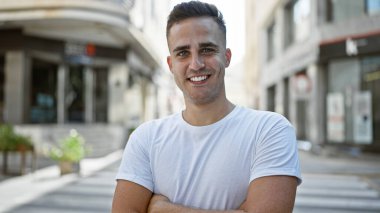 Gülümseyen genç İspanyol bir adam güvenli bir şekilde şehir sokak ortamında duruyor, yaklaşılabilirlik ve günlük tarz yayıyor..