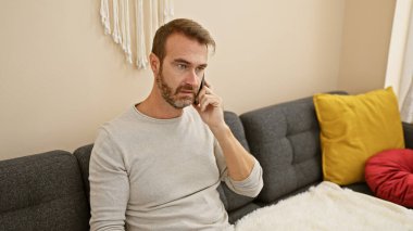 Düşünceli, orta yaşlı, sakallı, günlük gömlekli bir adam gri bir koltukta oturmuş, rahat bir oturma odasında telefonla konuşuyor..