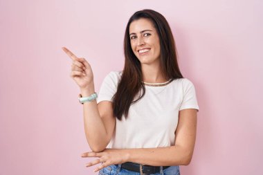 Young brunette woman standing over pink background with a big smile on face, pointing with hand finger to the side looking at the camera. 