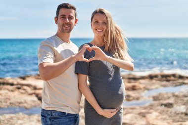 Kadın ve erkek bebek bekliyor. Deniz kenarında kalp jesti yapıyorlar.
