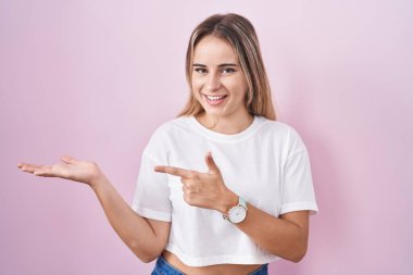 Young blonde woman standing over pink background amazed and smiling to the camera while presenting with hand and pointing with finger. 