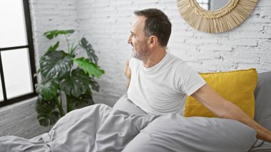 Olgun, kendine güvenen, gri saçlı, rahat bir gülümseme takınan, yatak odasında rahat bir yatakta mutlu bir şekilde sabahtan zevk alan bir adam.