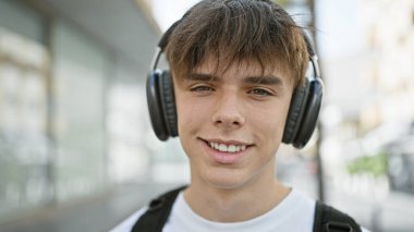 Genç Avrupalı sarışın bir adam açık havada kulaklıklarıyla gülümsüyor kalabalık bir şehir ortamında.