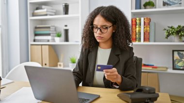 Genç ve güzel İspanyol iş kadını. Ofiste dizüstü bilgisayar ve kredi kartı kullanıyor.