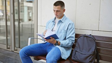 İspanyol bir adam kampüs bankında kitap okuyor, sırt çantası yanında, gençlik ve çalışkanlık yayıyor..