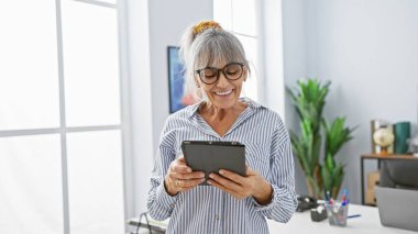 Ofis ortamında tablet kullanan gri saçlı, gülümseyen olgun bir kadın. Profesyonellik ve pozitiflik yayıyor..