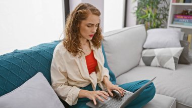 Oturma odasındaki kanepede oturan odaklanmış genç bir kadın dikkatle evdeki dizüstü bilgisayarını kullanıyor. Kafkasyalı güzel bir yetişkinin ciddi bir portresi. Cihazının üzerinde. Kapalı alandaki işine dalmış..