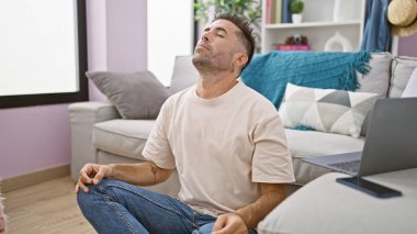 Çekici, genç İspanyol bir adam evinin rahat ortamında yoga egzersiziyle sağlığına kavuşuyor oturma odasında dinleniyor sakinliği kucaklıyor ve dengeyi buluyor..