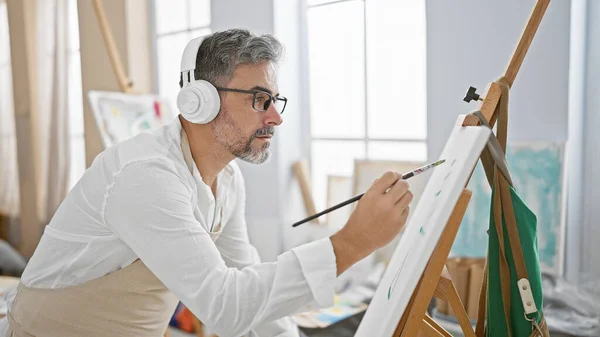 At the studio, a relaxed, young hispanic man with grey-haired beard, artist and student, drawn to music, drawing away on canvas, headphones on, bringing art to life indoors