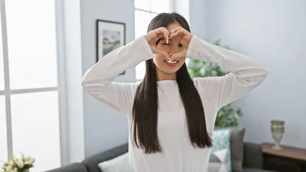 A cheerful asian woman making a heart shape with her hands in a bright living room