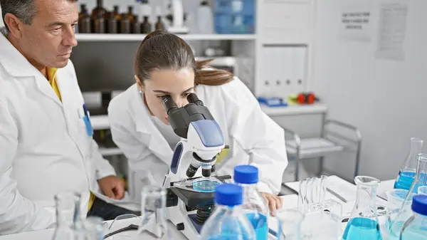 Hispanic man and woman as focused science partners, seriously working together in lab! zoom on chemistry experiment with microscope in bustling laboratory for medical research.