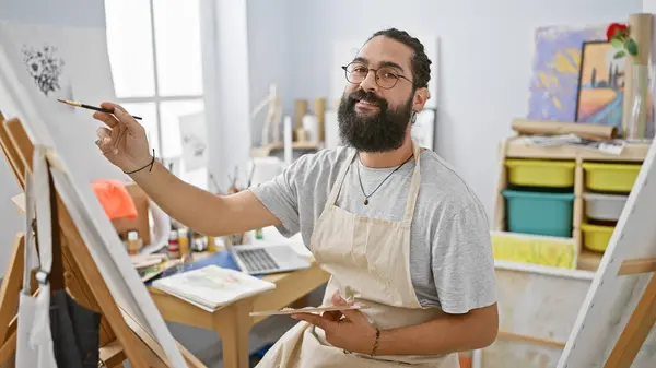 A bearded young man paints on a canvas in a bright art studio, embodying creativity and concentration.