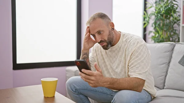 Surprised young man, sitting relaxed on sofa at home, drinking morning espresso from a mug, looking at his smartphone screen with amazed expression