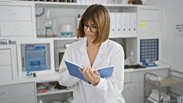 At the heart of science, young, beautiful hispanic woman scientist, relaxed yet concentrated, sitting at lab table, engrossed in a book\'s study, in the bustling center of medical breakthroughs