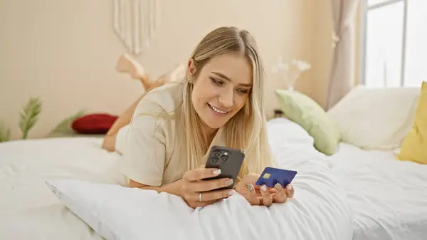 快乐的金发女子舒服地躺在床上 自信地享受着在舒适的卧室里用智能手机和信用卡进行网上购物的乐趣 — 图库照片