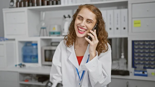 Smiling young woman scientist enthralled in marshaling medical marvels, animatedly talking on smartphone at her lab\'s work table