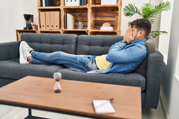 Young hispanic man patient stressed lying on sofa at psychology clinic