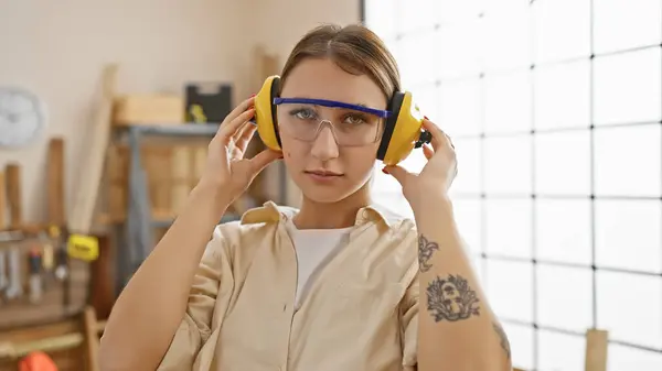 Woman in workshop wearing safety goggles and earmuffs looking focused while holding ear defenders.