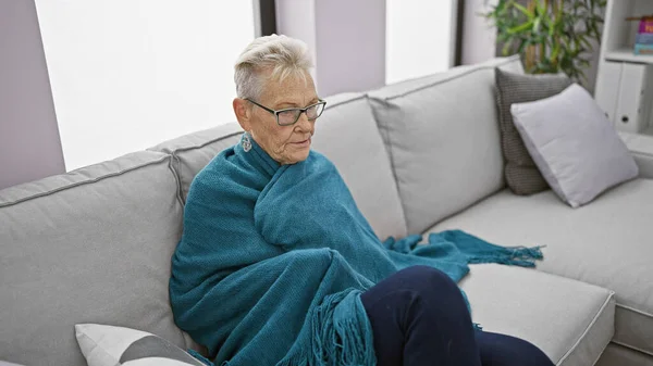 Senior grey-haired woman seeking warmth, snuggled in a blanket amid freezing temperature indoors, exhibiting signs of flu at her cozy home