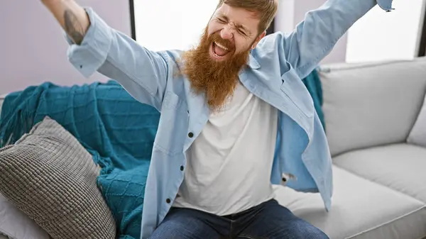 Joyous young redhead man, confidently celebrating a win, sitting on living room sofa, his handsome irish features reflecting the cheerful smile of victory