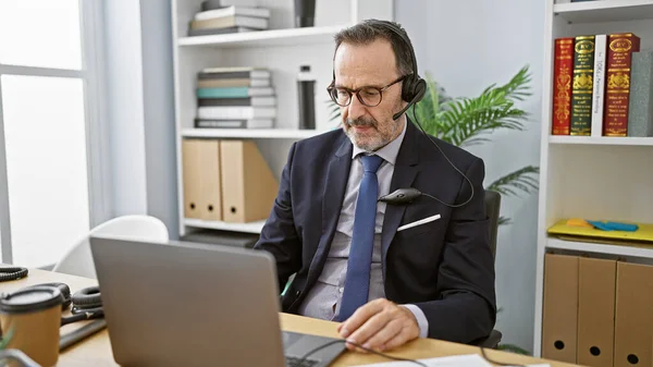 Hispanic middle-aged man with grey hair, focused on business work in the office, professionally speaking to a customer, providing aid through laptop. concentrated, successful worker using headset