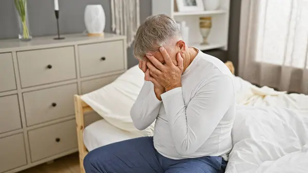 A distressed senior man sits holding his head in a modern bedroom, conveying worry, illness, or mental health issues.