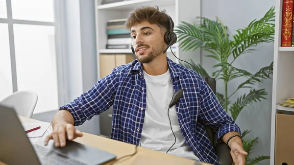 Beaming young arab man immersed in his business work, confidently rocking headphones, skilfully mastering his laptop in the bustling office environment.