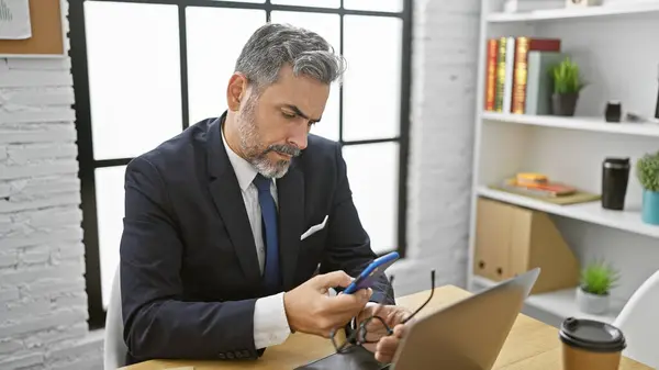 Overworked young hispanic man, grey-haired business worker, holding glasses whilst texting on smartphone in stressful office interior