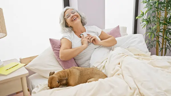 Uśmiechnięta Dojrzała Kobieta Leży Łóżku Psem Obok Niej Trzymając Telefon — Zdjęcie stockowe