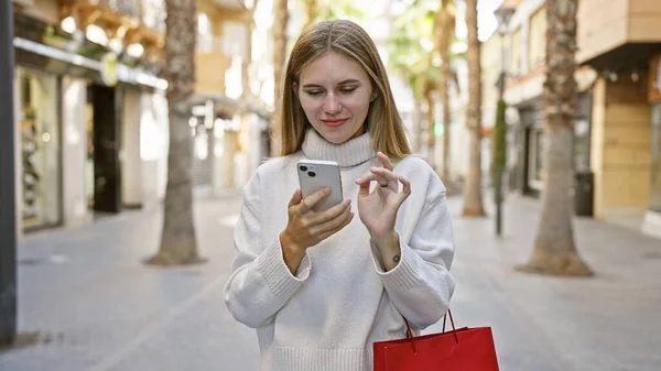 Blonde woman with blue eyes using smartphone on city street, holding shopping bag.