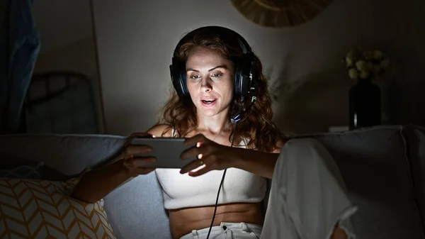 A young caucasian woman listens to music on headphones while using a smartphone, sitting on a couch indoors at night.