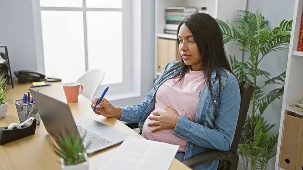 Pregnant young woman boss, taking notes while handling business over video call in office