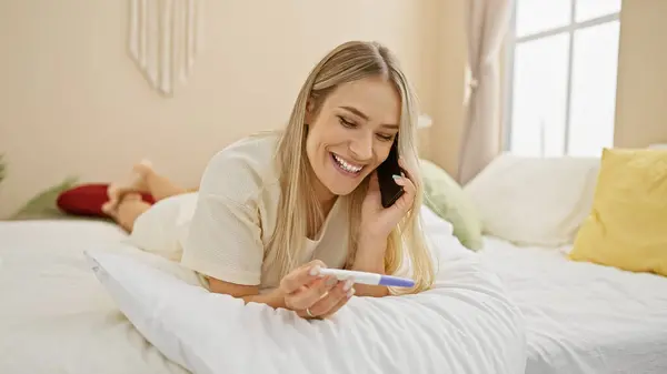 Candid shot of young, beautiful blonde woman, comfortably lying in bed in her cozy bedroom, talking playfully over the phone, her morning marked by a positive pregnancy test.