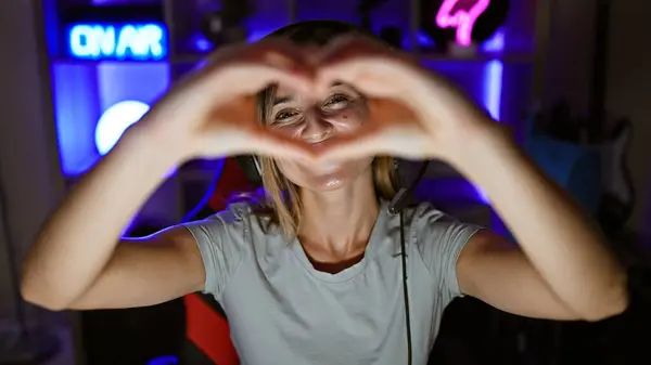 Confident young female gamer, a radiant blonde streamer, cheerfully flashes a heart symbol with her hands during a engaging night stream in her home-based gaming room