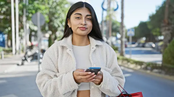 stock image A young indian woman with long hair using a smartphone on a sunny city street, holding a red shopping bag.