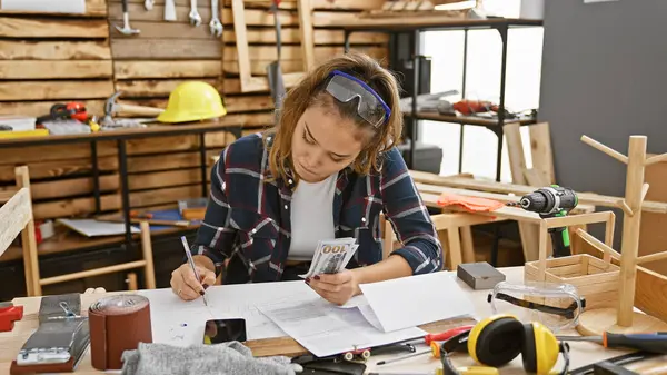 Young, beautiful hispanic woman carpenter counts dollars, taking business notes during woodwork session in indoor workshop