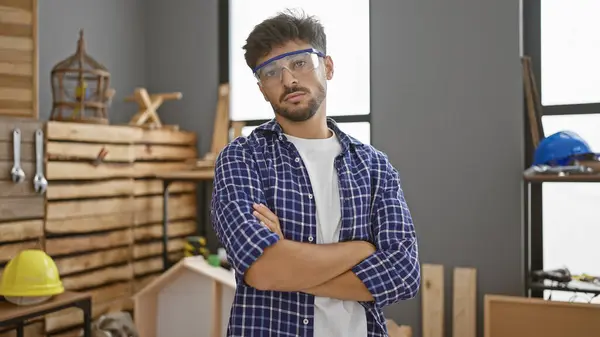 Serious-faced young arab carpenter with crossed arms standing formidably in his carpentry workshop, exemplifying the handsome adult male woodworker in the furniture industry
