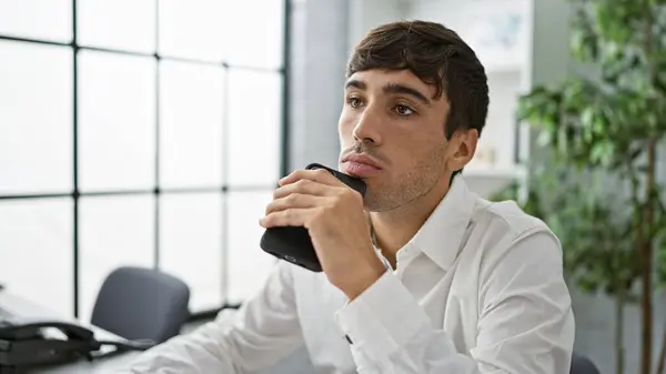 Handsome young hispanic man doubting work idea, seriously concentrating at his office job. portrait of an attractive business professional, a relaxed boss holding a smartphone, thinking indoors.